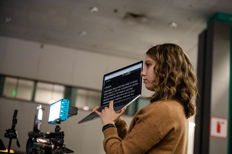 Sydney Handler holds up a computer with teleprompter words in front of a camera