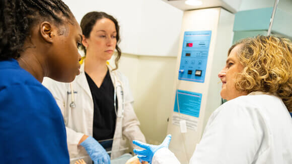 A professor instructs two nursing students over an infant manikin