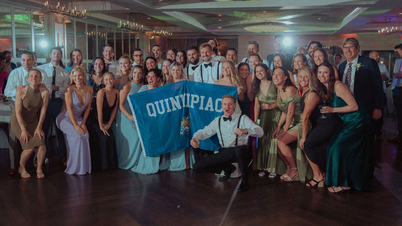 Emma and Spencer Mannion holding up a Quinnipiac flag at their wedding, surrounded by guests.