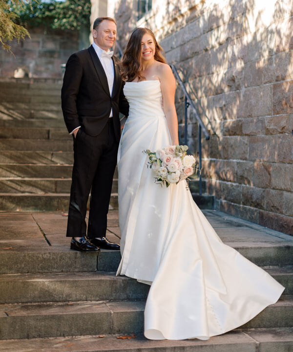 Jenn is wearing a long white wedding dress, standing on the steps with her husband Matt.