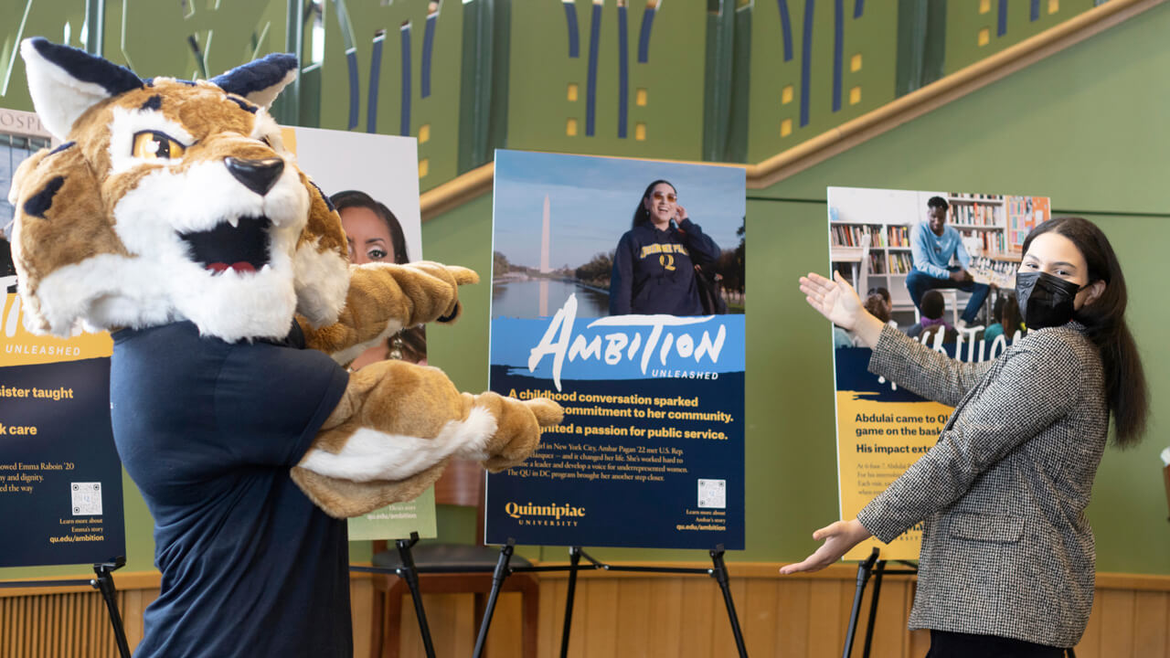 Ambar Pagan and Boomer the mascot celebrate in front of an Ambition Unleashed poster