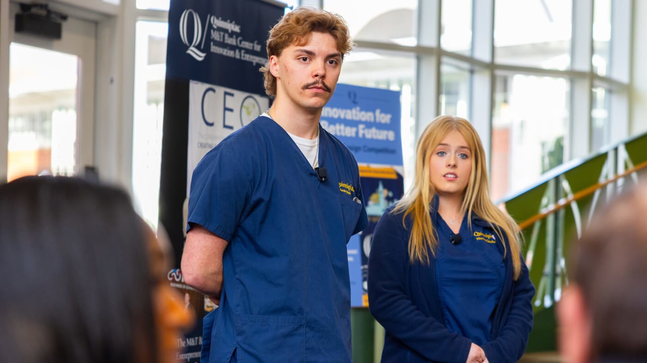 Two Quinnipiac students in navy blue scrubs stand at the front of a room presenting their business idea to a panel of judges during the pitch competition.