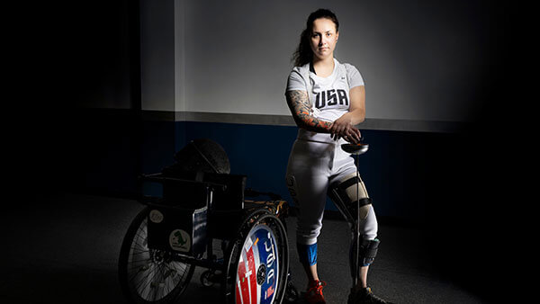 Victoria Isaacson stands in USA fencing gear next to their wheelchair.