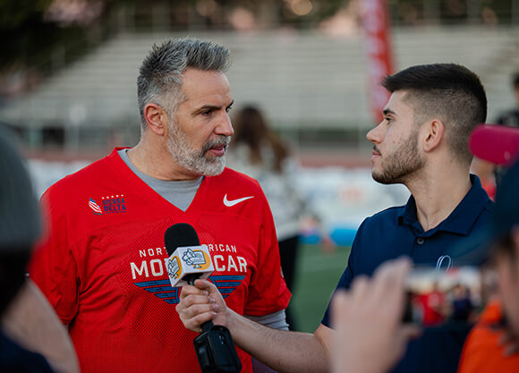 Santino Maione speaks with Kurt Warner on the sidelines of a football field