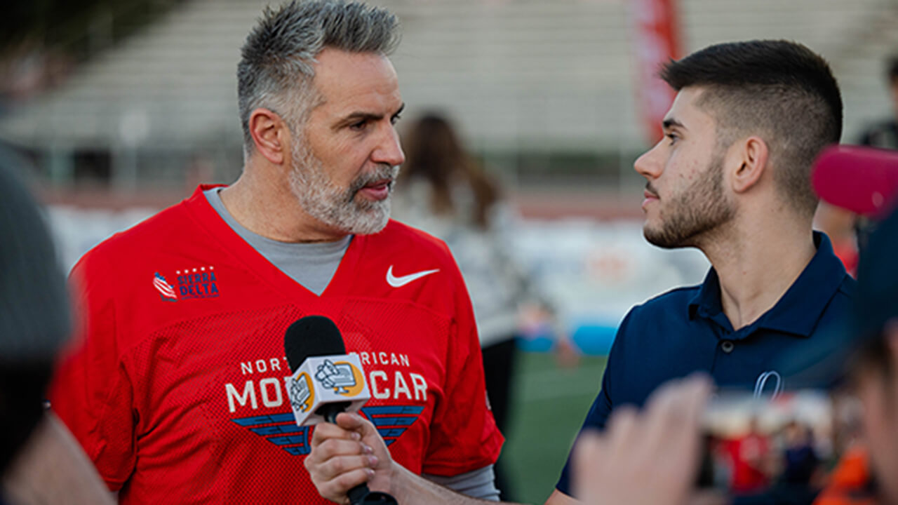 Santino Maione speaks with Kurt Warner on the sidelines of a football field