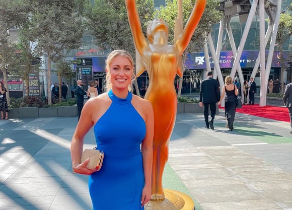 Natalie Pino stands in a blue dress outside with an Emmy's statue behind her.