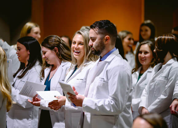 Physical therapy students smile in their white coats at a ceremony.