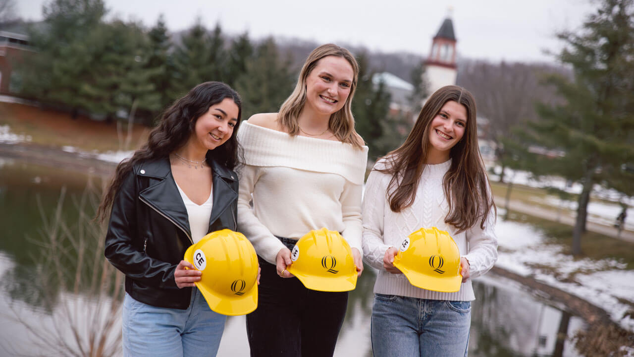 Three students smile holding hard hats outside in front of the library.
