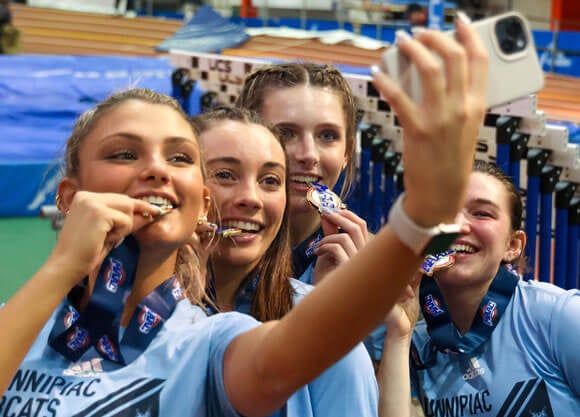 Four teammates on the women's track and field team smile and bite their medals in a selfie.