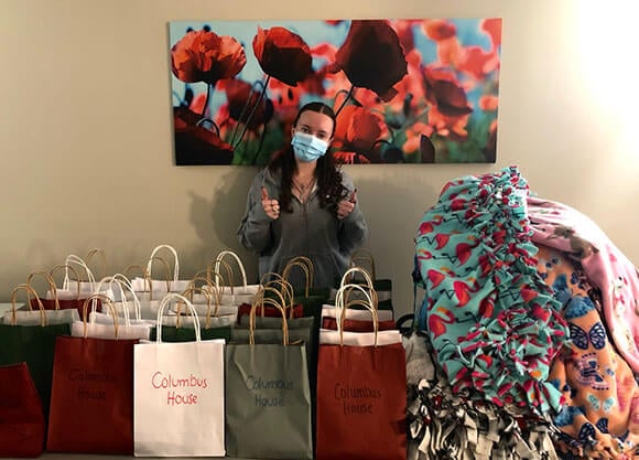 Student poses with gift bags labeled 'Columbus House' and has a pile of hand-tied fleece blankets