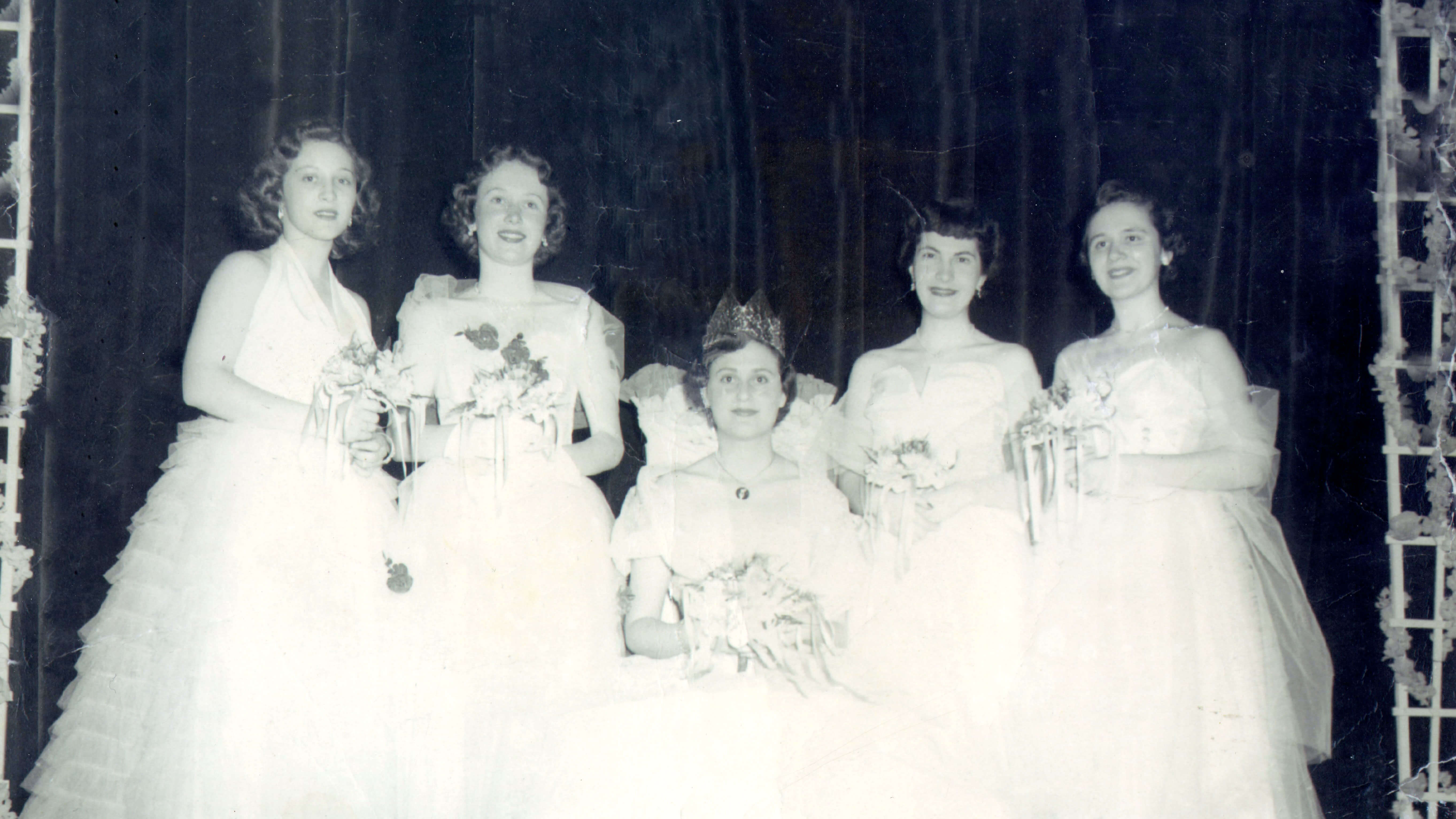 Five women smiling, dressed in white gowns while holding flowers