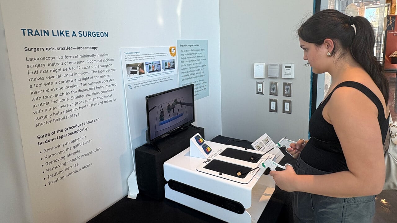 A female student stands in front of a museum exhibit of a medical device with the words "Train like a surgeon."