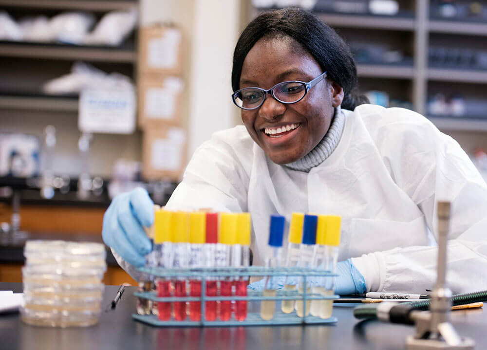 A student smiles and handles test tubes with gloved hands in the microbiology lab