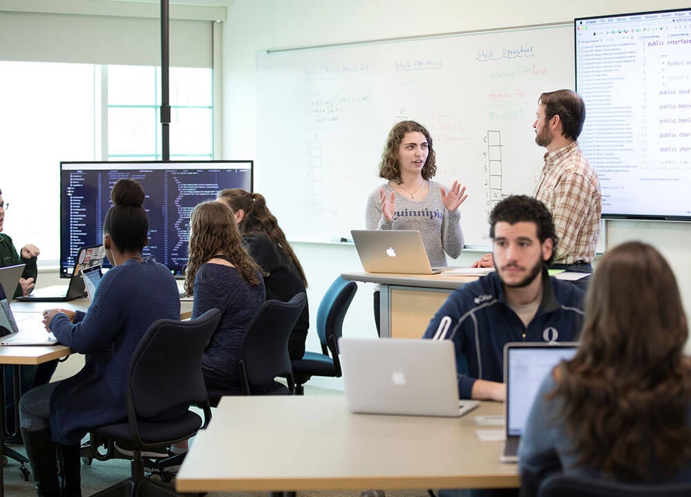 A group of students sit at a table and look at computer code on a large monitor in an engineering classroom