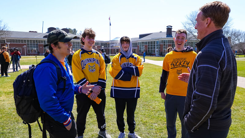 Fans in gold tee shirts and hockey jerseys speak with men's ice hockey coach Rand Pecknold on the quad