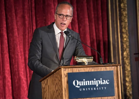 Lester Holt speaks at a podium in front of a red curtain at the Metropolitan Club in New York City