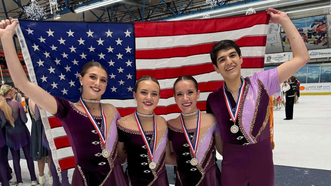 Four ice skaters in pink and purple costumes hold a large American flag on the ice.