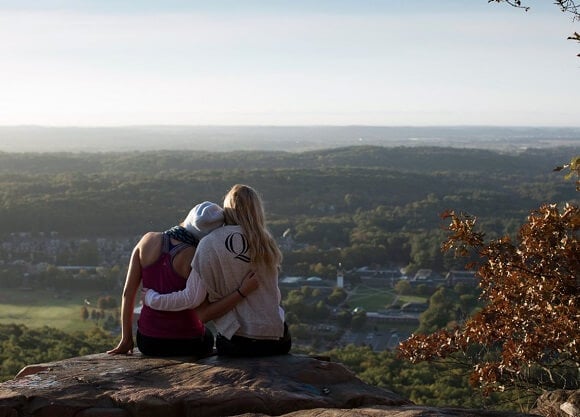 Two female students sit atop Sleeping Giant State Park overlooking Quinnipiac.