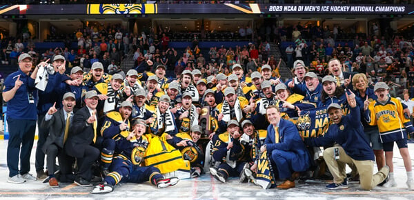 Men's ice hockey team and coaches cheer with the NCAA tournament trophy on the ice.