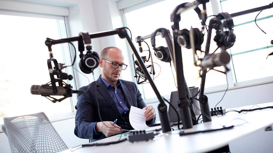 Professor David DesRoches sits behind a microphone with headphones on