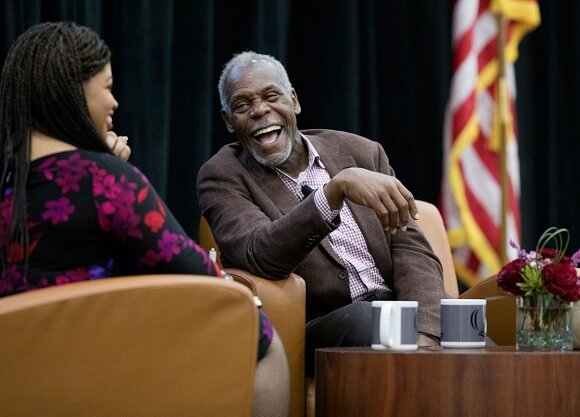 Danny Glover sits on a stage with an American flag, flowers and 'Q' mugs.
