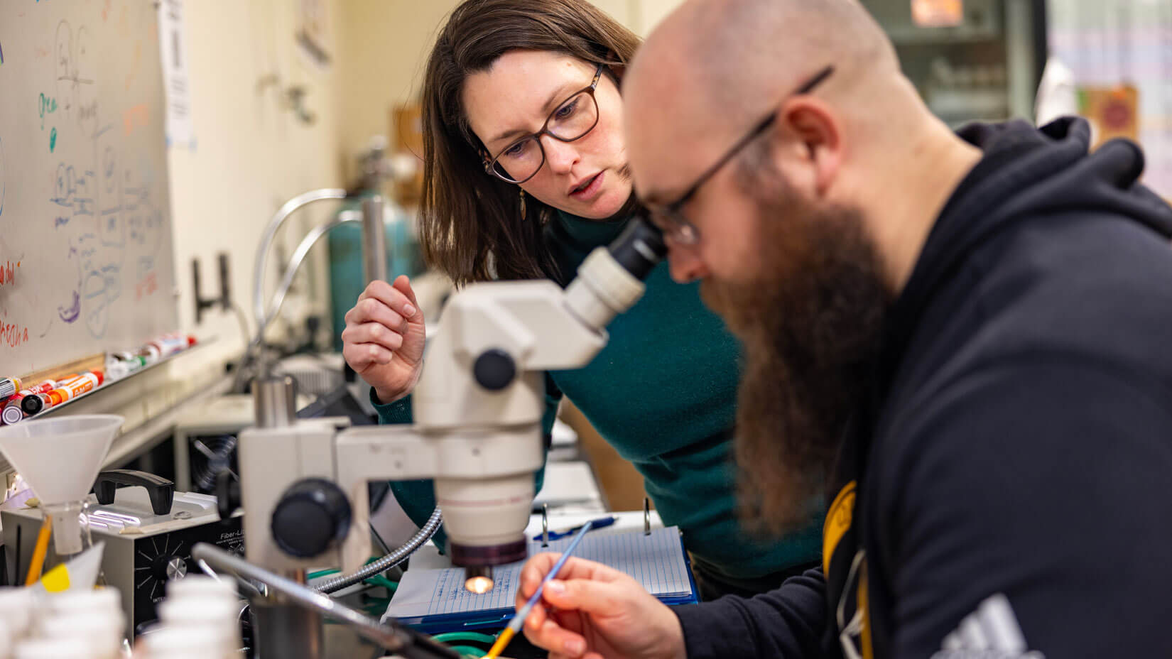 A student and professor in a lab examining a microscope slide together.