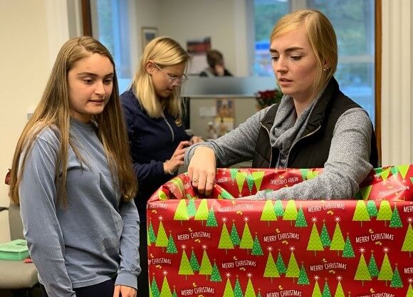 2 women wrap a box in red Christmas wrapping paper with green trees on it