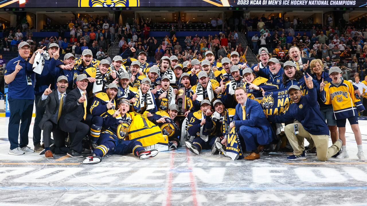 Men's ice hockey team and coaches cheer with the NCAA tournament trophy on the ice.