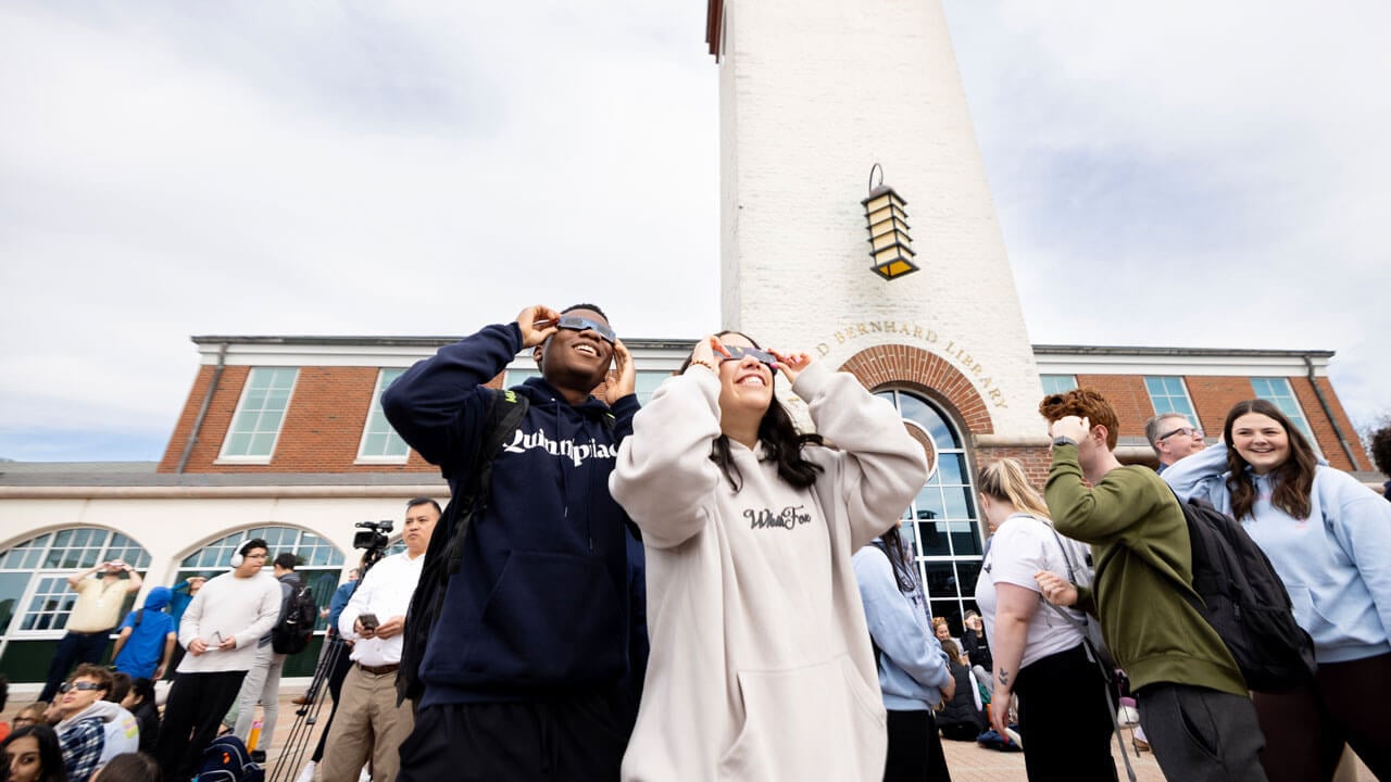 Two students hold up solar eclipse glasses to their eyes in a crowd in front of the library.