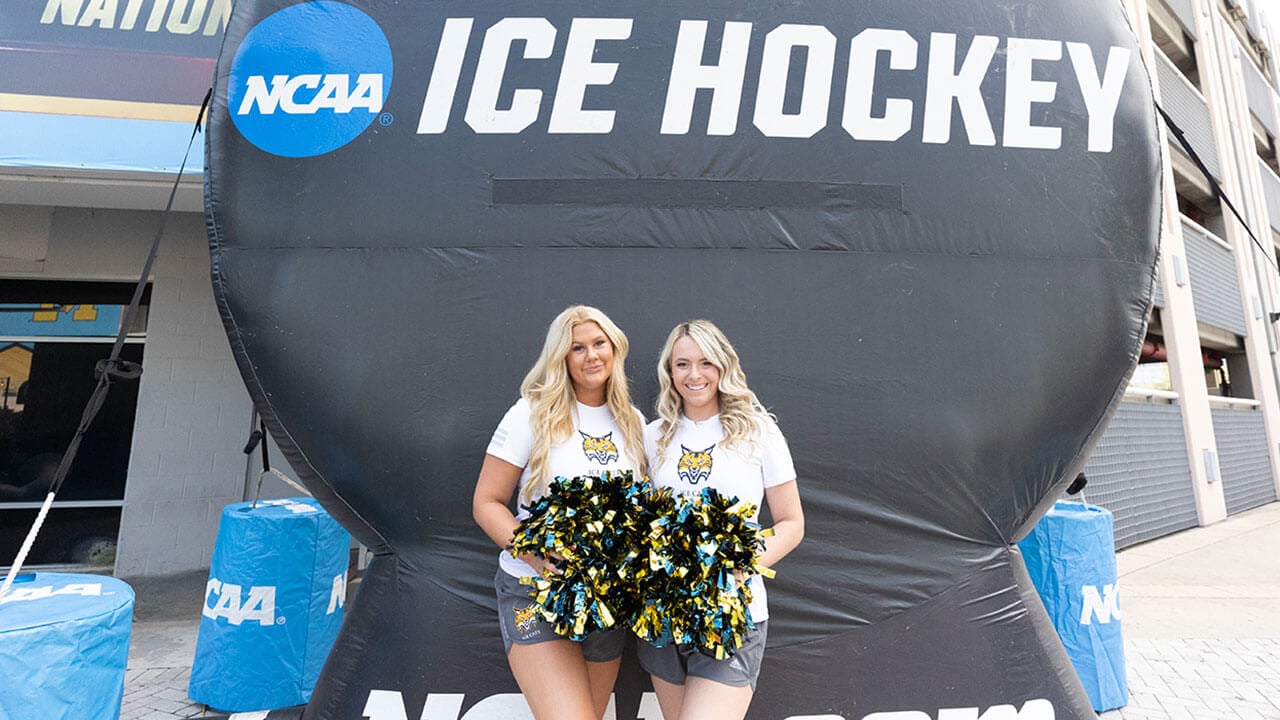 two ice cats pose with pom poms in front of an inflated NCAA hockey puck