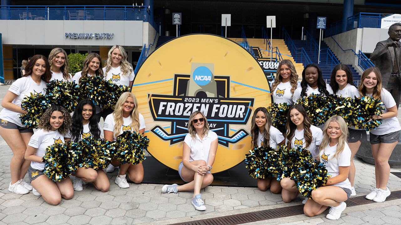 ice cats pose with their pom poms in front of puck shaped Frozen Four sign, their director sits on the ground in front
