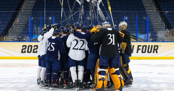 The men's ice hockey team huddles on the ice with their sticks in the air