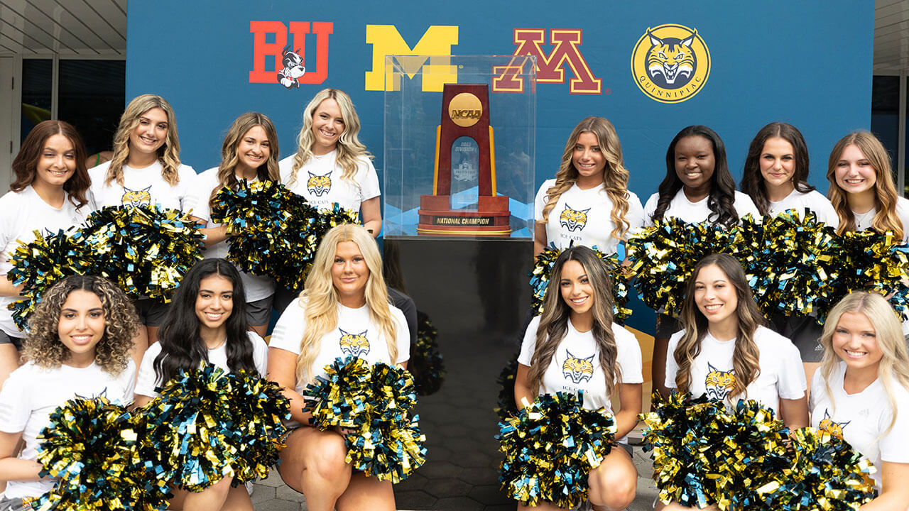 ice cats pose with pom poms and NCAA trophy in front of sign with all Frozen Four teams' logos