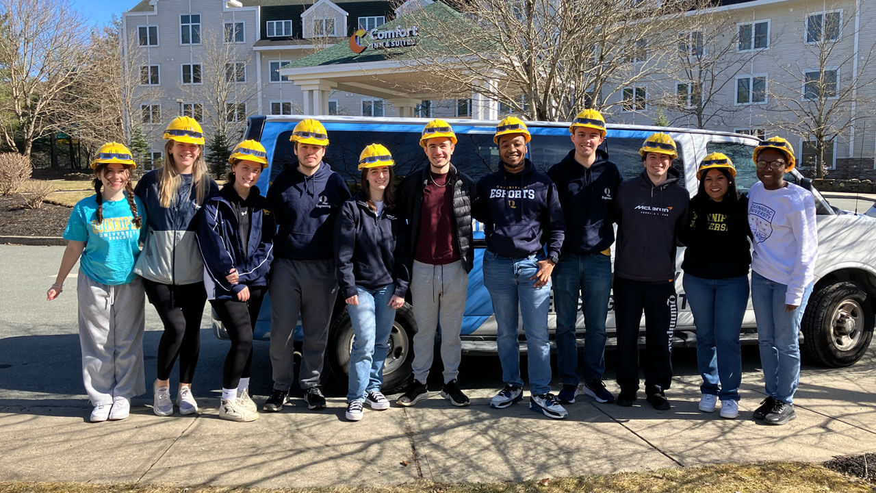 A group of students wear hard hats in front of a van.