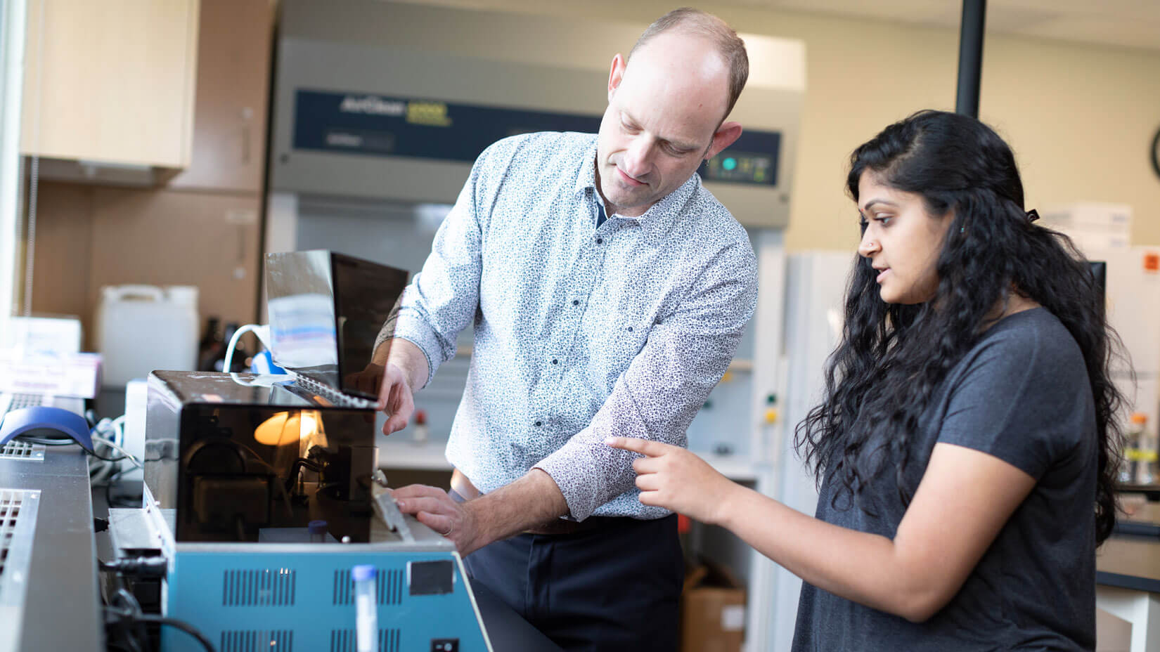 A biology professor guides a student to operate lab equipment.