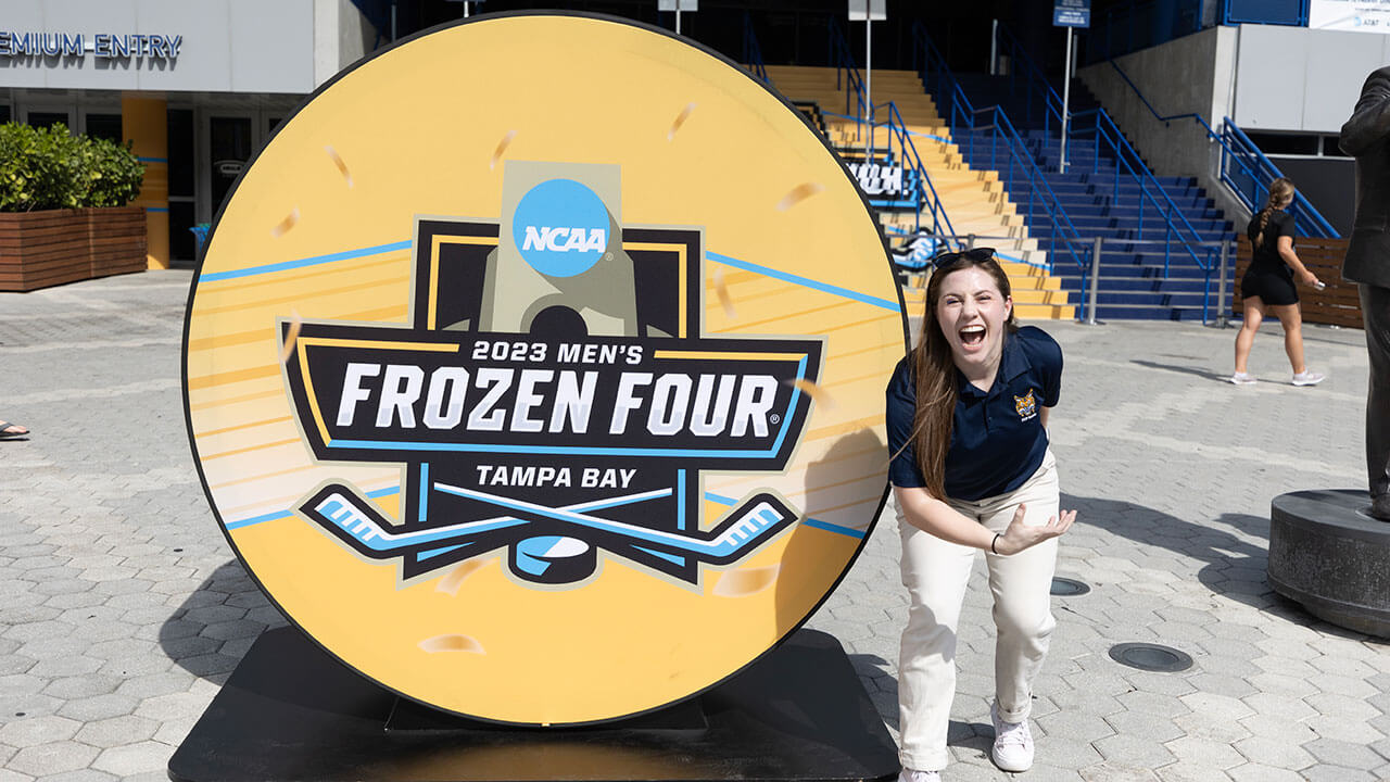 pep band member poses doing the bobcat roar with her arm beside Frozen Four sign