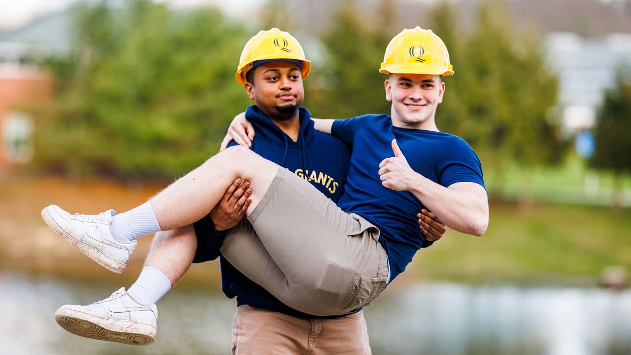 Engineer student carries other student in front of CCE pond while wearing hard hats