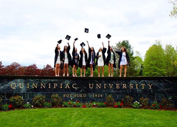 Students in graduation caps and gowns standing on the Quinnipiac University sign throwing caps in the air