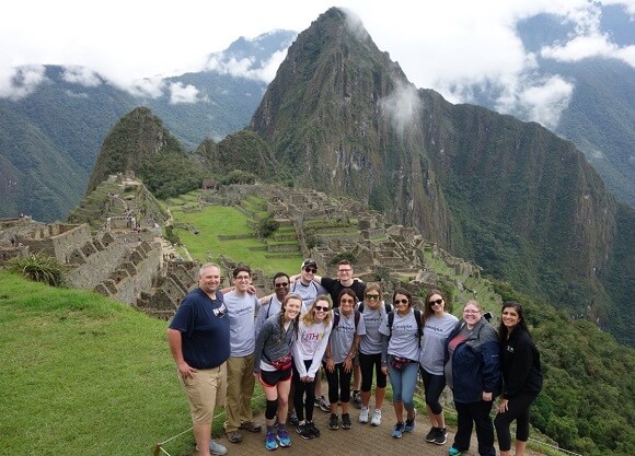 A group of students poses at the peak of a temple in Peru with clouds and mountains in the background