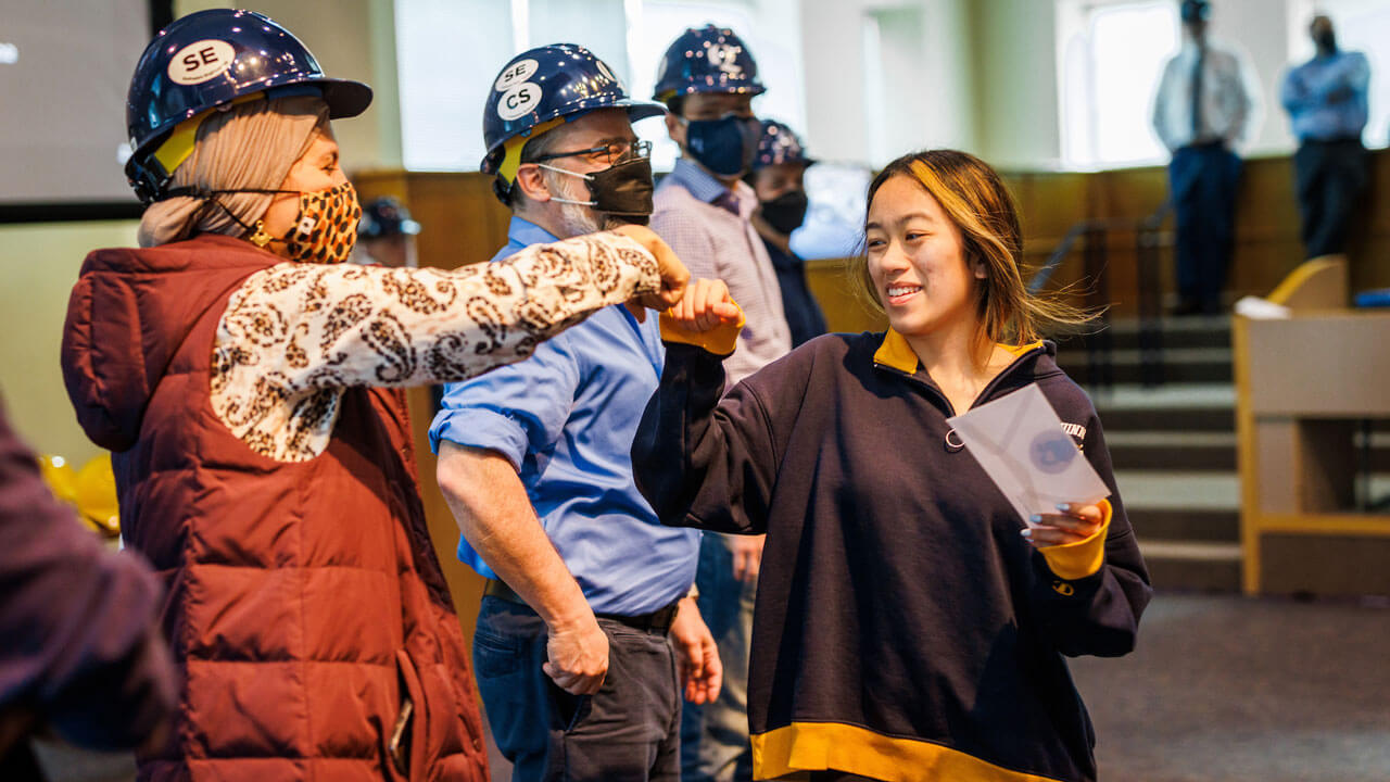 Girl fist bumps professor wearing navy blue hard hat in front of audience at hard hat ceremony