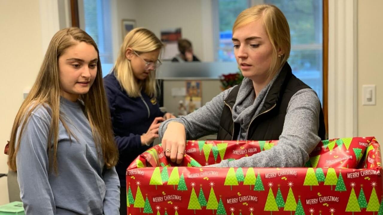 2 women wrap a box in red Christmas wrapping paper with green trees on it