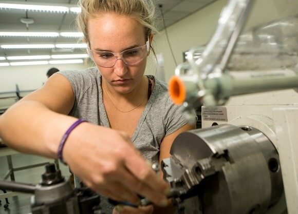 A female student wears safety goggles and works on a machine