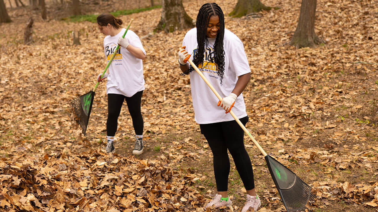 two female students rake to clear a path surrounded by trees