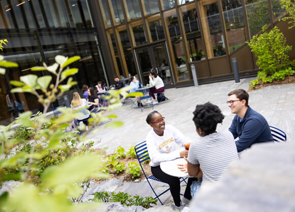 Small groups of students talk with each other at tables in the RecWell outdoor courtyard