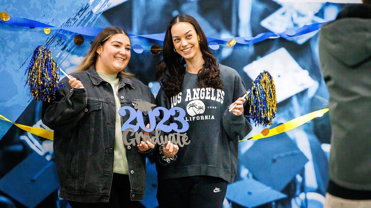 two female students with dark brown hair holding gold and navy blue shiny pom poms and a sign that says 2023 graduates