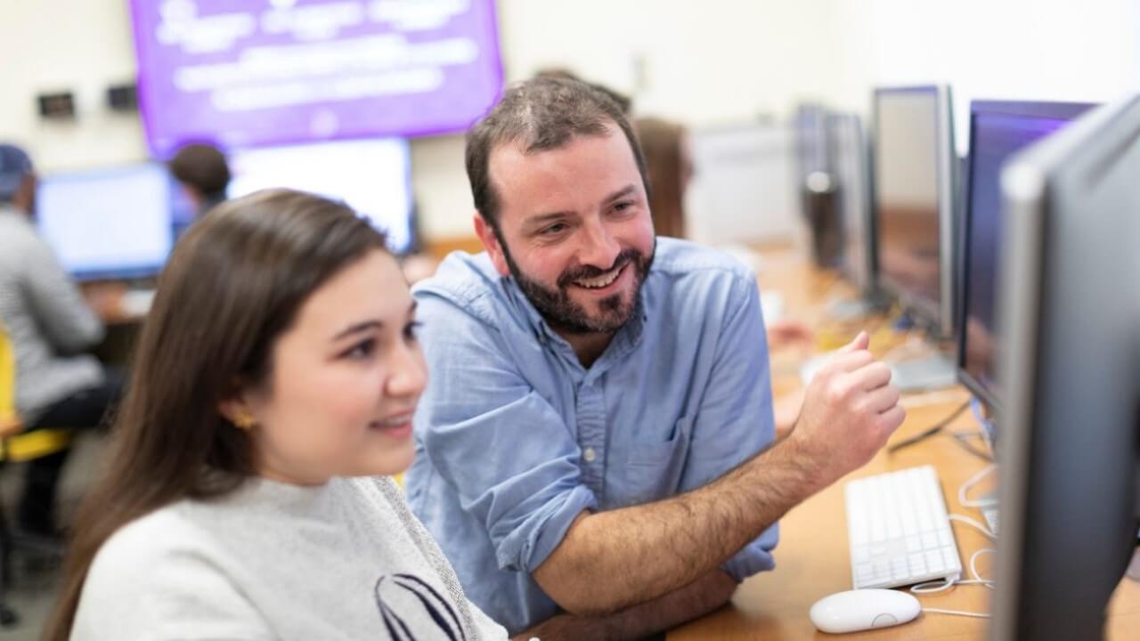 Jonah Warren sits at a table helping a female student with a video game project on a computer