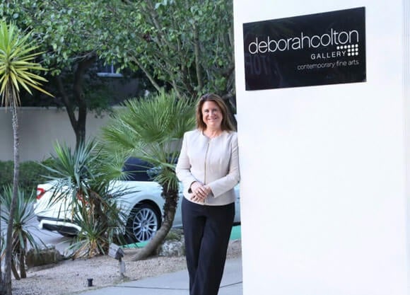 Deborah Colton leaning on a wall with a sign above her "Deborah Colton Gallery: contemporary fine arts"