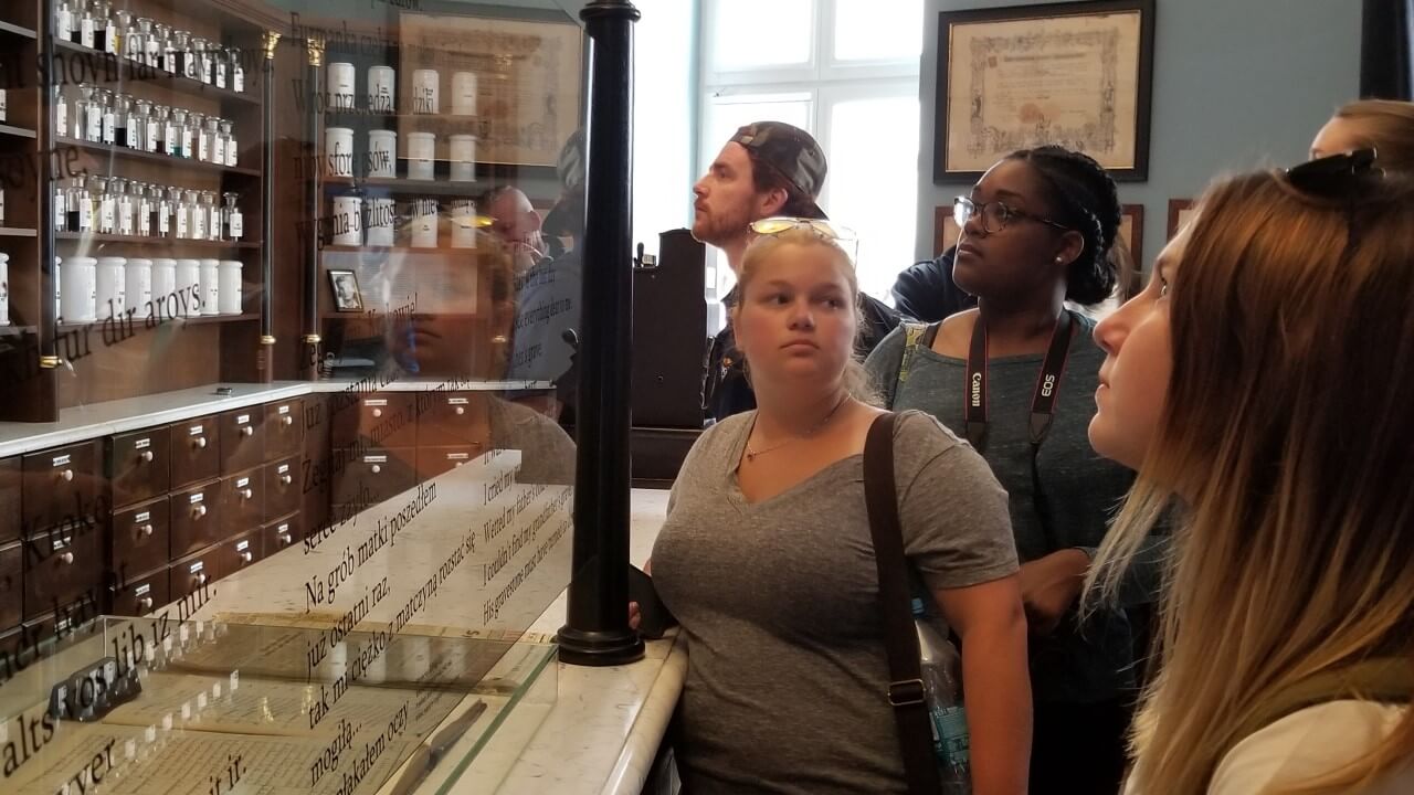 Students stand in a pharmacy from the 1940s looking at displays of glass containers.