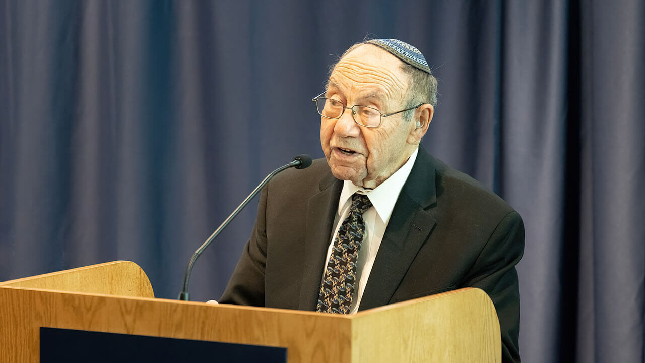 rabbi lazowski speaks behind a podium. he wears a navy blue and white yamaka