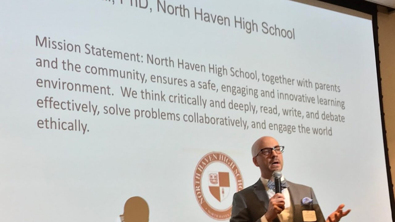 North Haven High School principal Russ Dallai gestures while holding a microphone in front of a screen.